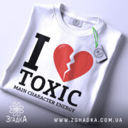 Футболка токсік енергія, білого кольору, складена на поверхні, з верхнього ракурсу, фон світлий, принт з червоним розбитим серцем та чорним текстом 'I LOVE TOXIC MAIN CHARACTER ENERGY', на комірці є етикетка.