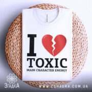 Футболка токсік енергія, білого кольору, складена на плетеній кошику, фон світлий, принт з червоним розбитим серцем та чорним текстом 'I LOVE TOXIC MAIN CHARACTER ENERGY'.