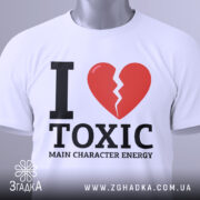 Футболка токсік енергія, білого кольору, складена на поверхні, з верхнього ракурсу, фон світлий, принт з червоним розбитим серцем та чорним текстом 'I LOVE TOXIC MAIN CHARACTER ENERGY', на комірці є етикетка.