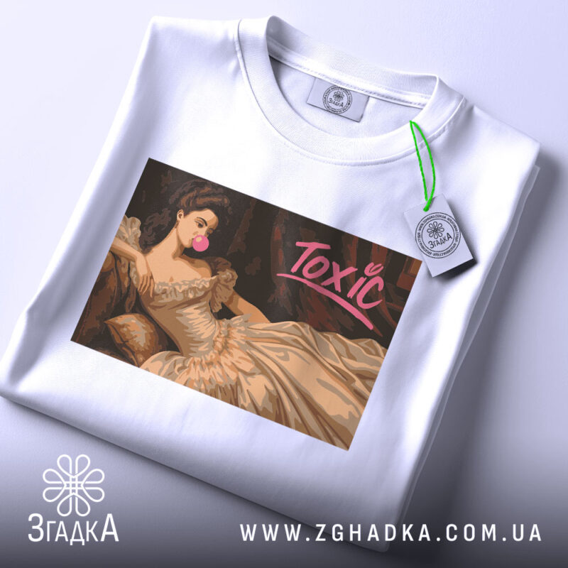 Футболка токсік білого кольору, складена на поверхні, з принтом, що зображує жінку в елегантній сукні, яка жує жувальну гумку. На футболці видно етикетку з зеленим шнурком. Принт має яскравий напис 'Toxic'.