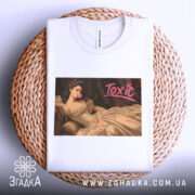 Футболка токсік білого кольору, складена на плетеній підставці, з принтом, що зображує жінку в елегантній сукні, яка жує жувальну гумку. Принт має яскравий напис 'Toxic'.