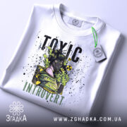 Футболка токсичний інтроверт, білого кольору, зображена на столі з верхнього ракурсу, фон світло-сірий, принт з дівчиною в протигазі, текст 'TOXIC INTRUVERT' в чорному та зеленому кольорах, на футболці є етикетка з зеленим шнурком.