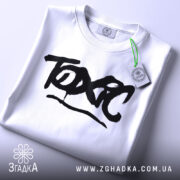 Футболка токсичний подарунок, біла, складена на столі, фон світло-сірий, принт чорний з написом 'TOXIC', на комірці є етикетка.