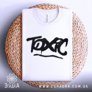 Футболка токсичний подарунок, біла, складена на плетеній тарілці, фон світло-сірий, принт чорний з написом 'TOXIC' у стилі графіті.