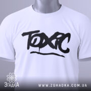 Футболка токсичний подарунок, біла, складена на столі, фон світло-сірий, принт чорний з написом 'TOXIC', на комірці є етикетка.