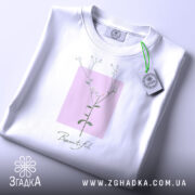 Футболка тонка ботаніка білого кольору, складена на поверхні, зображена з верхнього ракурсу, фон світло-сірий, принт з квітковим мотивом на рожевому квадраті з написом 'Beautiful' під ним, на футболці є етикетка з зеленим шнурком.