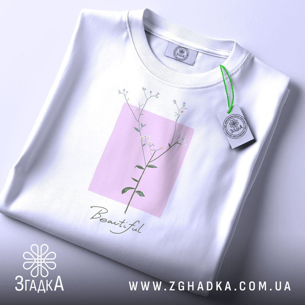 Футболка тонка ботаніка білого кольору, складена на поверхні, зображена з верхнього ракурсу, фон світло-сірий, принт з квітковим мотивом на рожевому квадраті з написом 'Beautiful' під ним, на футболці є етикетка з зеленим шнурком.