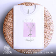 Футболка тонка ботаніка білого кольору, складена на плетеній кошику, зображена з верхнього ракурсу, фон світло-сірий, принт з квітковим мотивом на рожевому квадраті з написом 'Beautiful' під ним.