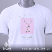 Футболка тонка ботаніка білого кольору, складена на поверхні, зображена з верхнього ракурсу, фон світло-сірий, принт з квітковим мотивом на рожевому квадраті з написом 'Beautiful' під ним, на футболці є етикетка з зеленим шнурком.