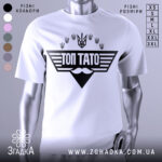 Футболка топ тато білого кольору, зображена з близького ракурсу на манекені, фон світлий, принт чорного кольору з написом 'ТОП ТАТО' та символікою України.