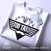 Футболка топ тато білого кольору, складена на поверхні, з видимим етикеткою, фон світлий, принт чорного кольору з написом 'ТОП ТАТО' та символікою України.