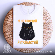 Футболка товстий пухнастий, біла, зображена зверху на плетеній основі, на фоні світло-сірого кольору, принт з чорним котом з жовтими очима та текстом "Я не товстий, я пухнастий" у жовтому кольорі.
