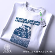 Футболка трактор у полі, біла, складена на столі, фон світлий, принт з трактором та текстом синього кольору, на футболці є етикетка.