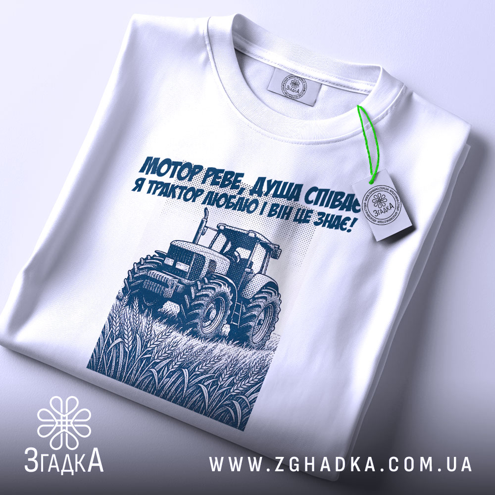 Футболка трактор у полі, біла, складена на столі, фон світлий, принт з трактором та текстом синього кольору, на футболці є етикетка.