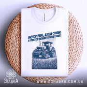 Футболка трактор у полі, біла, складена на плетеній основі, фон світлий, принт з трактором та текстом синього кольору, видно етикетку на комірці.