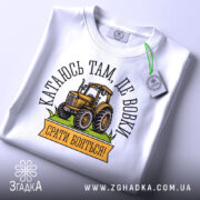 Футболка тракторист білого кольору, складена на столі, фон світло-сірий, принт з жовтим трактором та написом "Катаюсь там, де вовки, срати бояться!", на комірці видна етикетка.