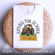 Футболка тракторист білого кольору, складена на плетеній основі, фон світло-сірий, принт з жовтим трактором та написом "Катаюсь там, де вовки, срати бояться!".