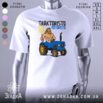 Футболка Трактористо мотористо білого кольору, зображена на манекені, з принтом, що зображає м'язистого чоловіка з довгим волоссям, який тримає тризуб, стоячи поруч з синім трактором. Фон нейтральний, з легким градієнтом.