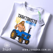 Футболка Трактористо мотористо білого кольору, складена на столі, з принтом, що зображає м'язистого чоловіка з тризубом і синім трактором. На футболці є етикетка з зеленим шнурком. Фон світлий, з легким відблиском.