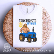 Футболка Трактористо мотористо білого кольору, складена на плетеному кошику, з принтом, що зображає м'язистого чоловіка з тризубом і синім трактором. Фон світлий, з текстурою плетеного кошика.