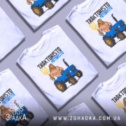 Футболка Трактористо мотористо білого кольору, зображена на манекені, з принтом, що зображає м'язистого чоловіка з довгим волоссям, який тримає тризуб, стоячи поруч з синім трактором. Фон нейтральний, з легким градієнтом.