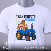 Футболка Трактористо мотористо білого кольору, складена на столі, з принтом, що зображає м'язистого чоловіка з тризубом і синім трактором. На футболці є етикетка з зеленим шнурком. Фон світлий, з легким відблиском.