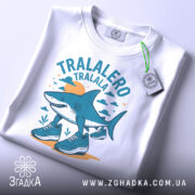 Футболка Tralalero Tralala білого кольору, зображена зверху на столі, фон світло-сірий. Принт з акулами у спортивному взутті, оточений хмарами та сонцем, виконаний у блакитних та жовтих тонах, з етикеткою на шнурку.