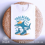 Футболка Tralalero Tralala білого кольору, зображена зверху на плетеній кошику, фон світло-сірий. Принт з акулами у спортивному взутті, оточений хмарами та сонцем, виконаний у блакитних та жовтих тонах.