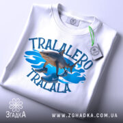 Футболка tralalero tralala, білого кольору, зображена зверху, складена на поверхні, фон світлий, принт з акулами та кросівками на блакитних хвилях, з етикеткою.