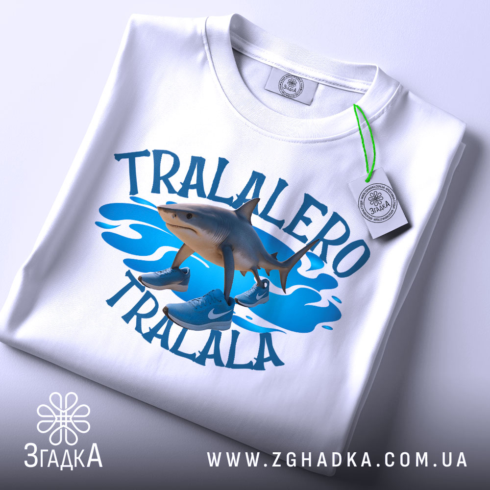 Футболка tralalero tralala, білого кольору, зображена зверху, складена на поверхні, фон світлий, принт з акулами та кросівками на блакитних хвилях, з етикеткою.
