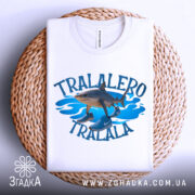 Футболка tralalero tralala, білого кольору, зображена зверху, складена на плетеній основі, фон світлий, принт з акулами та кросівками на блакитних хвилях.