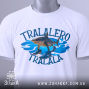 Футболка tralalero tralala, білого кольору, зображена зверху, складена на поверхні, фон світлий, принт з акулами та кросівками на блакитних хвилях, з етикеткою.