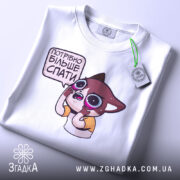 Футболка треба більше спати. Складена біла футболка з принтом мультяшного персонажа, що виглядає здивованим, з великими очима. На футболці є етикетка з зеленим шнурком. Фон світло-сірий, що надає акцент на футболку.