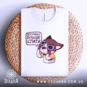 Футболка треба більше спати. Складена біла футболка з принтом мультяшного персонажа, що виглядає здивованим, на плетеній кошику. Фон світло-сірий, що підкреслює деталі принта.