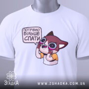 Футболка треба більше спати. Складена біла футболка з принтом мультяшного персонажа, що виглядає здивованим, з великими очима. На футболці є етикетка з зеленим шнурком. Фон світло-сірий, що надає акцент на футболку.
