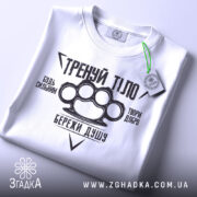 Футболка тренуй тіло, білого кольору, складена на поверхні, фон світло-сірий, принт чорного кольору з написами та зображенням кулака, на футболці є етикетка з зеленим шнурком.