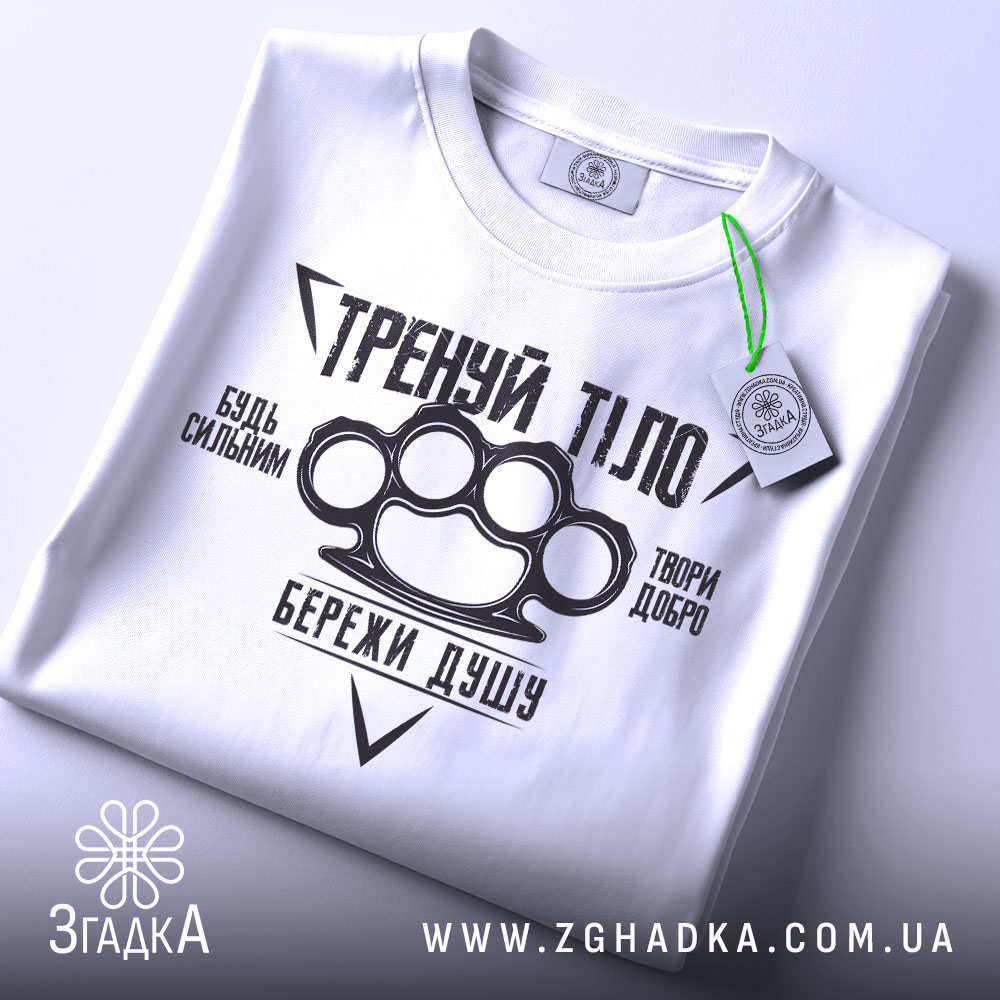 Футболка тренуй тіло, білого кольору, складена на поверхні, фон світло-сірий, принт чорного кольору з написами та зображенням кулака, на футболці є етикетка з зеленим шнурком.