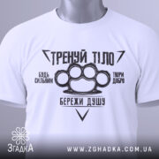 Футболка тренуй тіло, білого кольору, складена на поверхні, фон світло-сірий, принт чорного кольору з написами та зображенням кулака, на футболці є етикетка з зеленим шнурком.