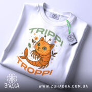 Футболка Trippi Troppi білого кольору, зображена зверху, на ній видно принт з оранжевим котом, оточеним рослинами, напис 'TRIPPI TROPPi' у зеленому та оранжевому кольорах, на футболці також є етикетка з зеленим шнурком.