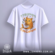 Футболка Trippi Troppi білого кольору, зображена з фронтального ракурсу, підвішена на вішалці, фон світло-сірий, принт з оранжевим котом, що має антенки, оточений квітами та написом 'TRIPPI TROPPi' у зеленому та оранжевому кольорах.