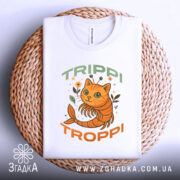 Футболка Trippi Troppi білого кольору, зображена зверху на плетеній основі, принт з оранжевим котом, оточеним рослинами, напис 'TRIPPI TROPPi' у зеленому та оранжевому кольорах, видно етикетку на комірці.