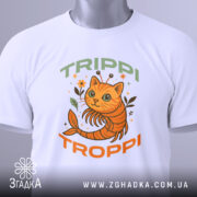 Футболка Trippi Troppi білого кольору, зображена зверху, на ній видно принт з оранжевим котом, оточеним рослинами, напис 'TRIPPI TROPPi' у зеленому та оранжевому кольорах, на футболці також є етикетка з зеленим шнурком.