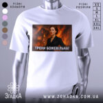 Футболка трохи божевільна, білого кольору, з прямим ракурсом, на фоні світло-сірого, з великим принтом, що зображує жінку в чорному костюмі на фоні палаючого будинку.