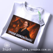 Футболка трохи божевільна, білого кольору, з верхнього ракурсу, на фоні світло-сірого, з великим принтом, що зображує жінку в чорному костюмі, а також з етикеткою на зеленому шнурку.