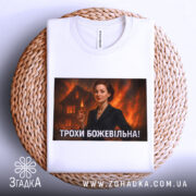 Футболка трохи божевільна, білого кольору, з верхнього ракурсу, на фоні плетеного кошика, з великим принтом, що зображує жінку в чорному костюмі на фоні палаючого будинку.