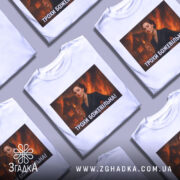 Футболка трохи божевільна, білого кольору, з прямим ракурсом, на фоні світло-сірого, з великим принтом, що зображує жінку в чорному костюмі на фоні палаючого будинку.