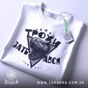 Футболка трохи за*бався, біла, зображена зверху, складена на поверхні, фон світло-сірий, принт з чорним малюнком голови динозавра та написом 'ТРОХИ ЗАБ'АВСЯ' у трикутнику, з зеленим ярликом.