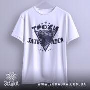 Футболка трохи за*бався, біла, зображена на вішалці, фон світло-сірий, принт з чорним малюнком голови динозавра та написом 'ТРОХИ ЗАБ'АВСЯ' у трикутнику з зірками.