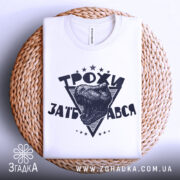 Футболка трохи за*бався, біла, зображена зверху, складена на плетеній основі, фон світло-сірий, принт з чорним малюнком голови динозавра та написом 'ТРОХИ ЗАБ'АВСЯ' у трикутнику.