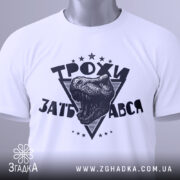 Футболка трохи за*бався, біла, зображена зверху, складена на поверхні, фон світло-сірий, принт з чорним малюнком голови динозавра та написом 'ТРОХИ ЗАБ'АВСЯ' у трикутнику, з зеленим ярликом.